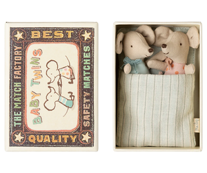 A set of two small toy mice designed to look like baby twins inside an open matchbox. The matchbox is adorned with illustrations and has a striped pattern inside.