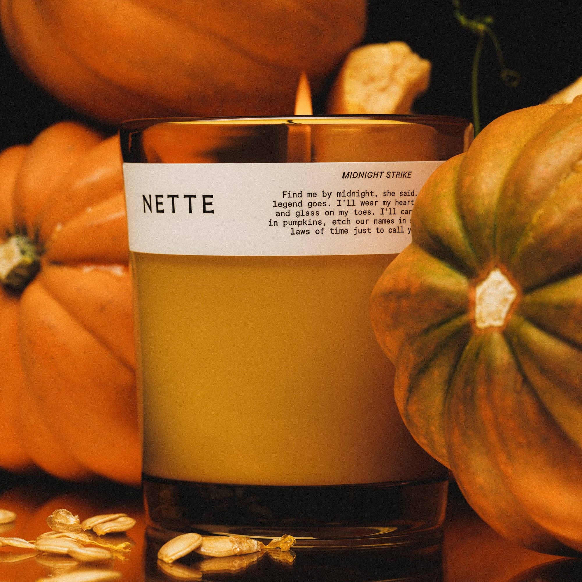 Candle labeled 'NETTE' surrounded by pumpkins on a reflective surface