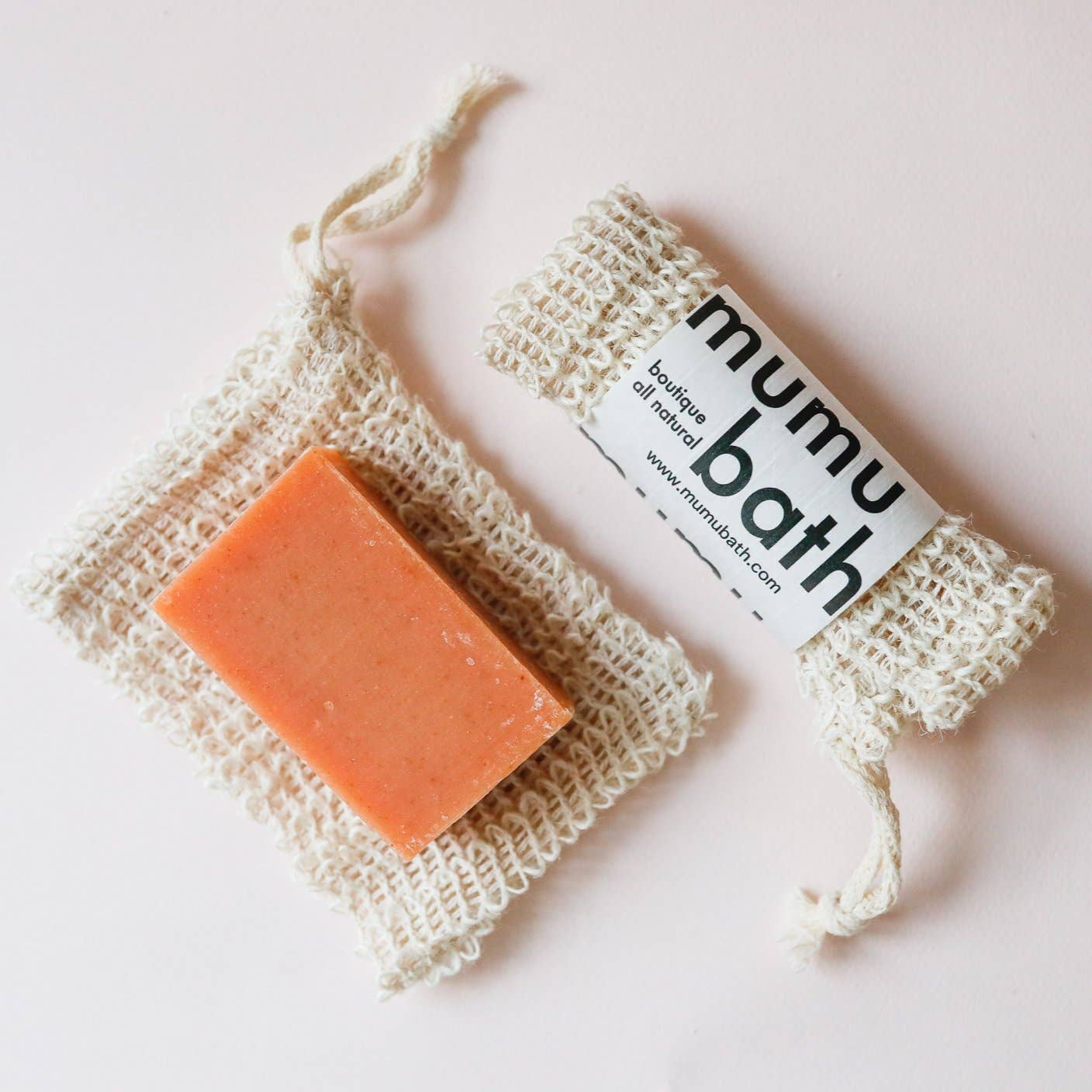 A bar of soap placed inside a beige, agave fiber soap saver pouch with the label 'MUMU BATH' on top, secured with a twine loop.