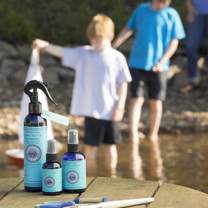 Three blue bottles with black sprayers on a wooden surface with children playing in the background.