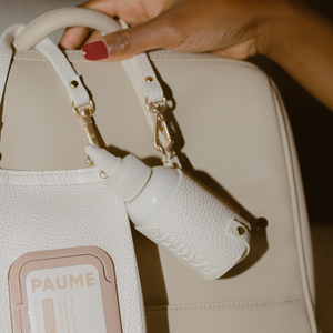 Close-up of a hand holding a beige handbag with a white water bottle holder, featuring the brand 'PAUME'.