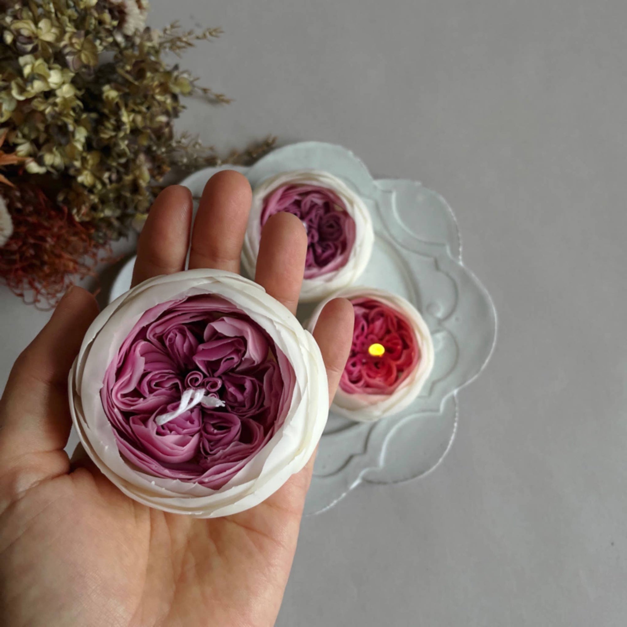 A hand holding a rose-shaped candle with layers of pink and white petals, placed on a white dish with more candles and dried flowers in the background.