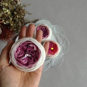 A hand holding a rose-shaped candle with layers of pink and white petals, placed on a white dish with more candles and dried flowers in the background.
