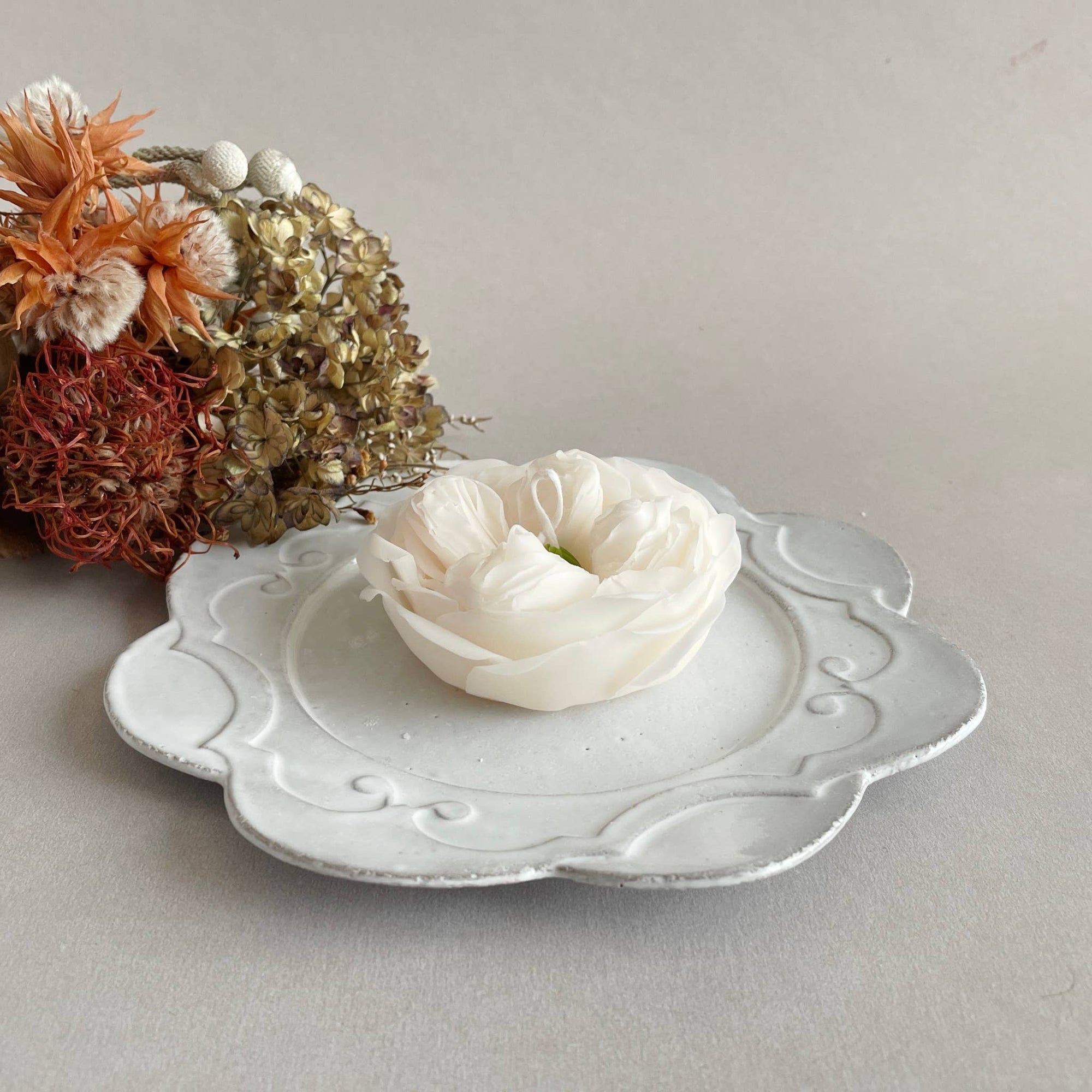 Decorative white dish with a small white flower on a light gray surface with dried flowers in the background.