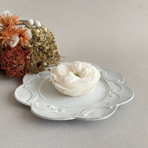 Decorative white dish with a small white flower on a light gray surface with dried flowers in the background.