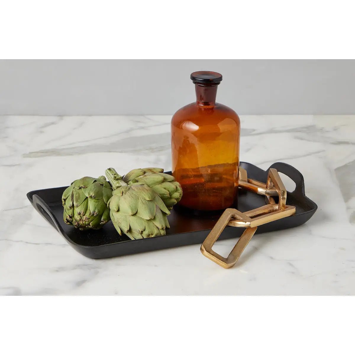 A black tray on a marble surface with three artichokes, an amber pharmacy bottle, and a gold link decoration. 