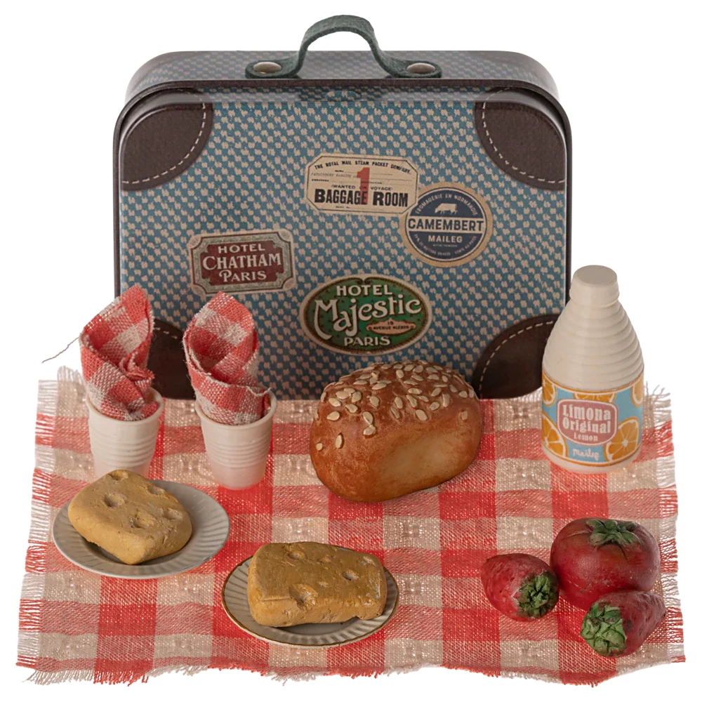 A small metal suitcase with a 'Maileg' brand tag, painted to look like a picnic basket, accompanied by toy food items like a pretend sandwich, cake, and drinks on a red and white checkered blanket.