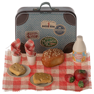 A small metal suitcase with a 'Maileg' brand tag, painted to look like a picnic basket, accompanied by toy food items like a pretend sandwich, cake, and drinks on a red and white checkered blanket.