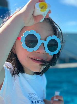 Child wearing blue sunglasses with flower designs, holding a tube of Play sunscreen, against a blurred outdoor background.