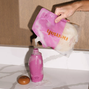 A person is holding a pink baby wash and shampoo refill bottle, which is designed to look like a milk carton. The bottle is being emptied into a white sink with a marble surface, alongside a brown egg.