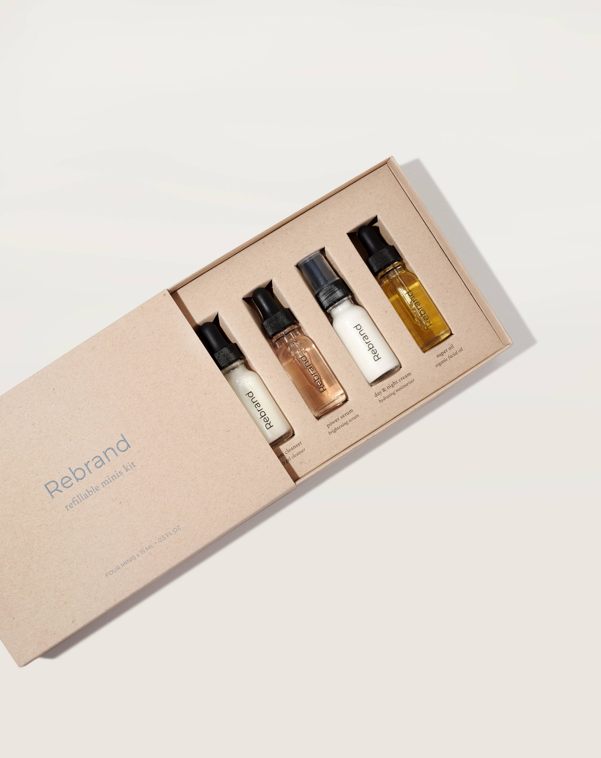 A collection of four 15 ML glass bottles with dropper caps, labeled Dream Cleanser, Power Serum, Day & Night Cream, and Super Oil, positioned in front of a brown cardboard box with the brand name Rebrand visible.