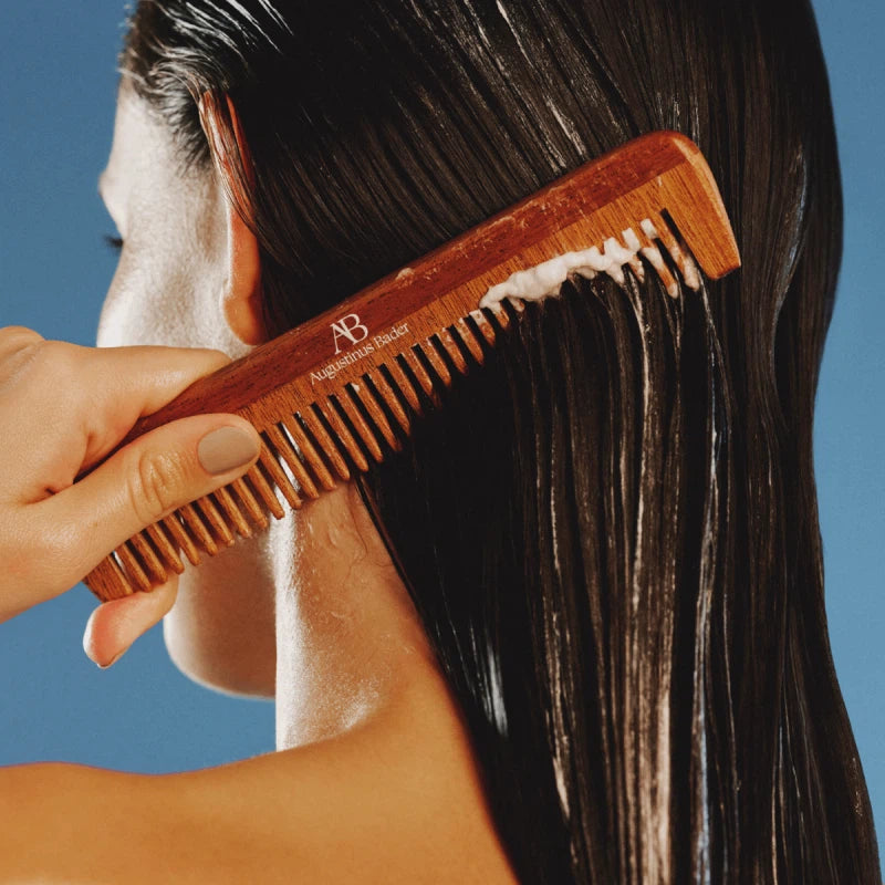 Person applying hair product to their hair with a wooden comb against a clear blue sky.