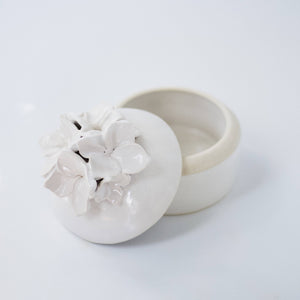 A cream-colored lidded jar with hand-sculpted flowers attached to the lid, placed on a white background.