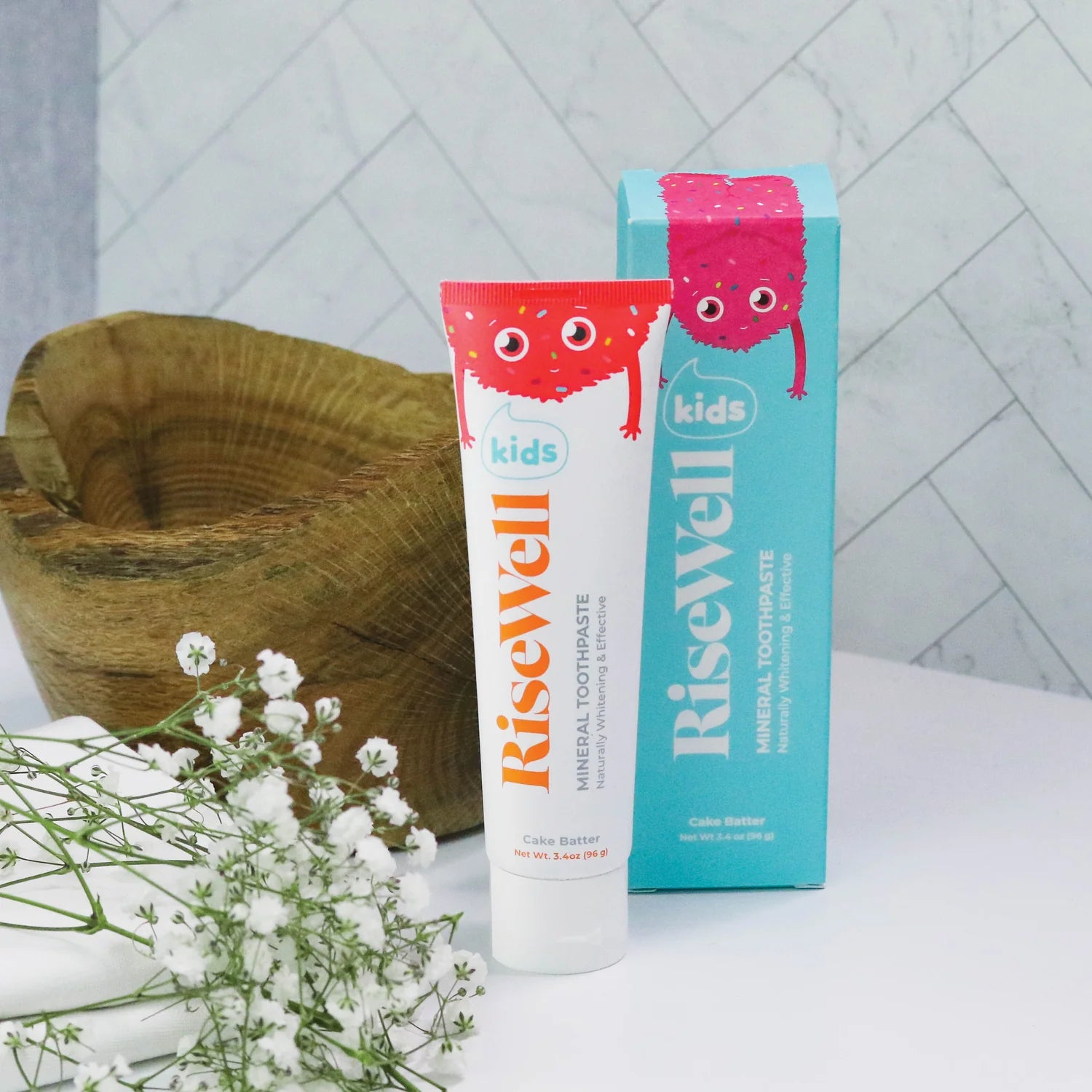RiseWell kids' toothpaste tube and packaging on a white surface with a wooden bowl and flowers in the background.