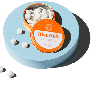 Container with orange lid featuring 'RiseWell' brand, filled with white mints on a white background.