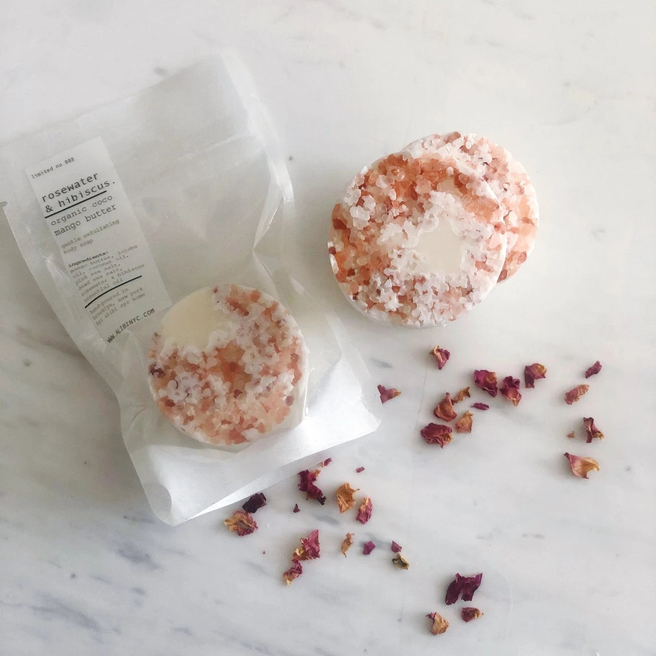 Three soaps with rose petals on a marble surface, accompanied by a tube of bath butter.