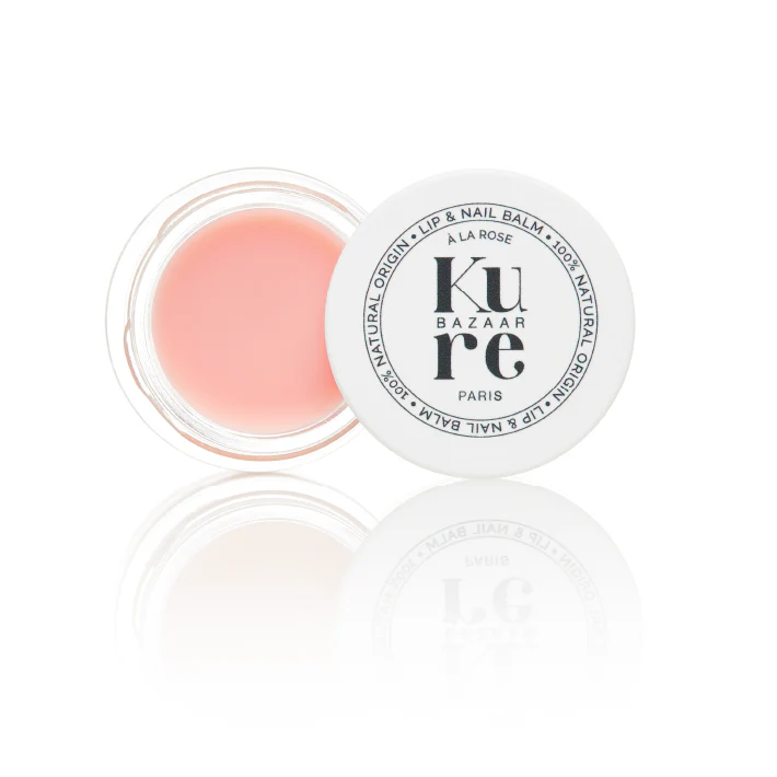 A pot of Kure Bazaar Lip & Nail Balm with the lid off, showing a pinkish-colored balm inside. The product is labeled with the brand name and the product name in black text on a white background.
