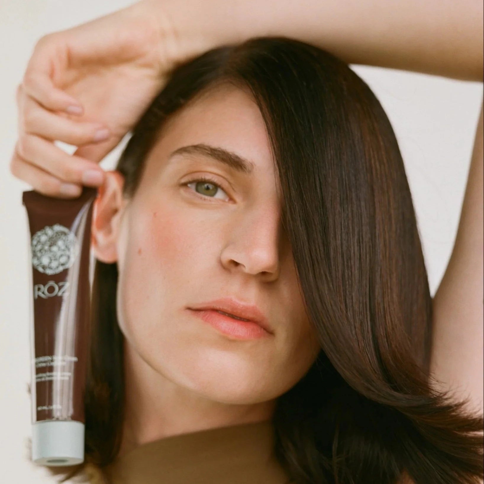 A woman with dark shoulder length hair holds a bottle of evergreen styling cream above her chest