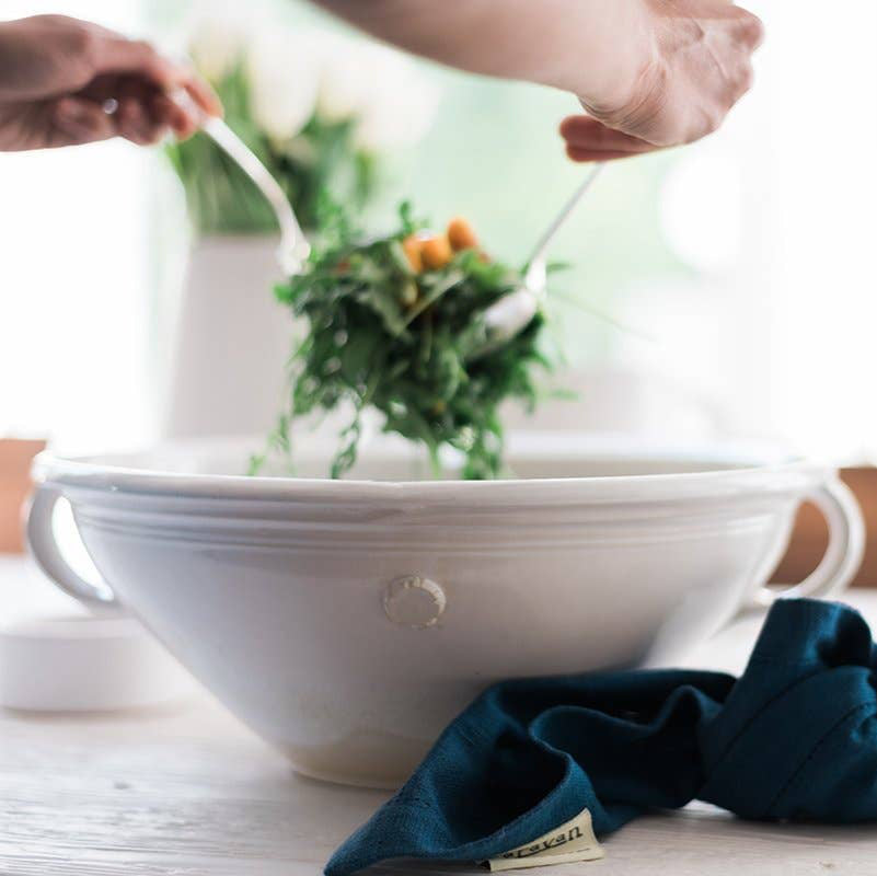 A white mixing bowl on a table, with a person scooping salad out of it. 