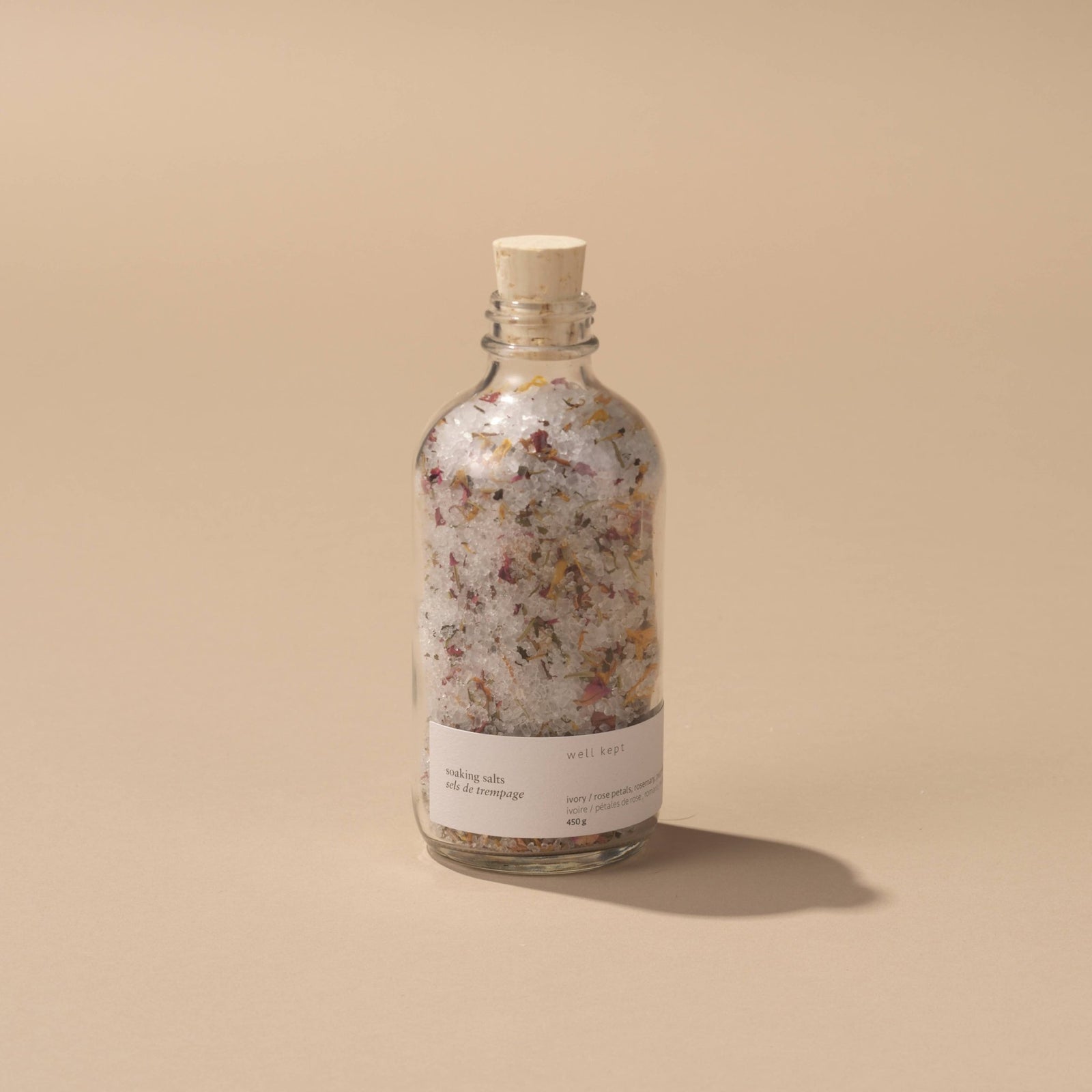 A clear glass bottle with a cork stopper, filled with pink and white floral bath salts. The label reads 'Well Kept' and indicates the size as 450g.