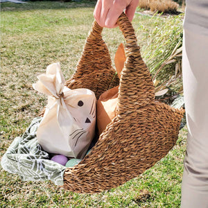 A person holding a Savar Handwoven Picnic Basket with stuffed animal bunnies and some greenery. 
