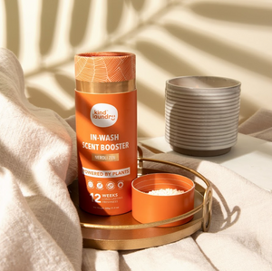 A bottle of Kind Laundry In Wash Scent Boosters placed on a gold tray next to a linen fabric, with a tropical background and palm leaves.