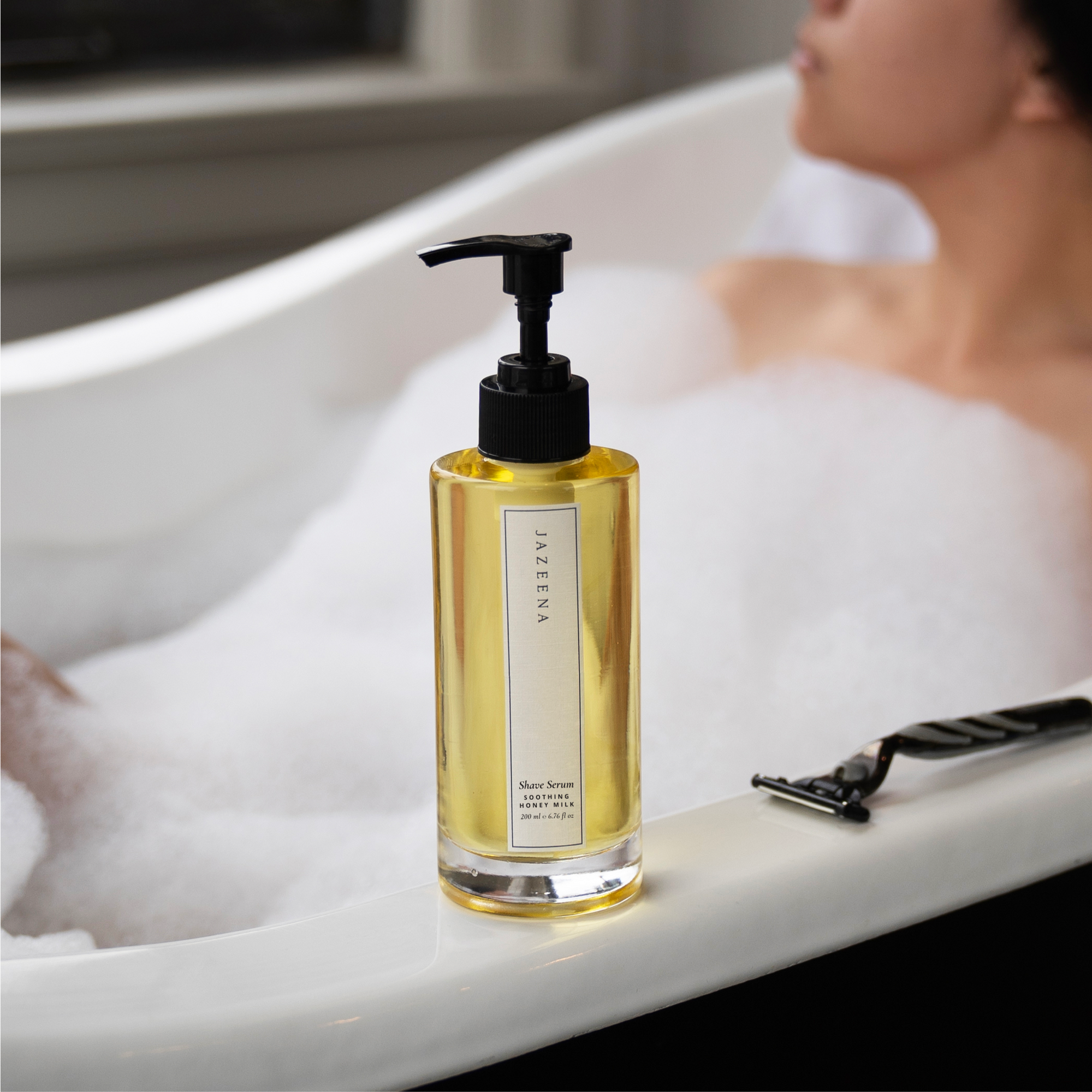 A bottle of Jazeena Shave Serum placed on the edge of a bathtub filled with bubbles, with a person relaxing in the background.
