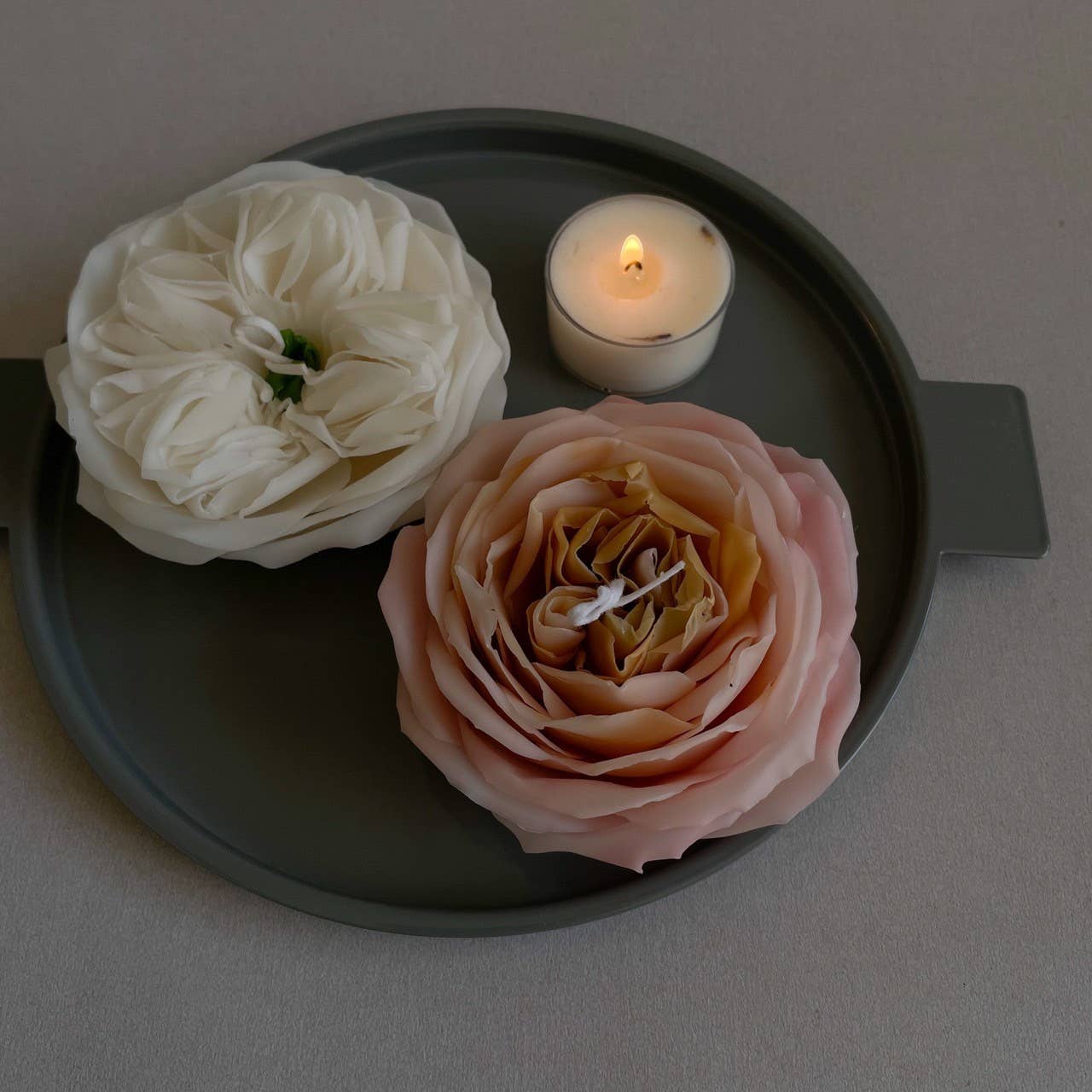 Two flowers and a lit candle on a dark gray tray against a light gray background