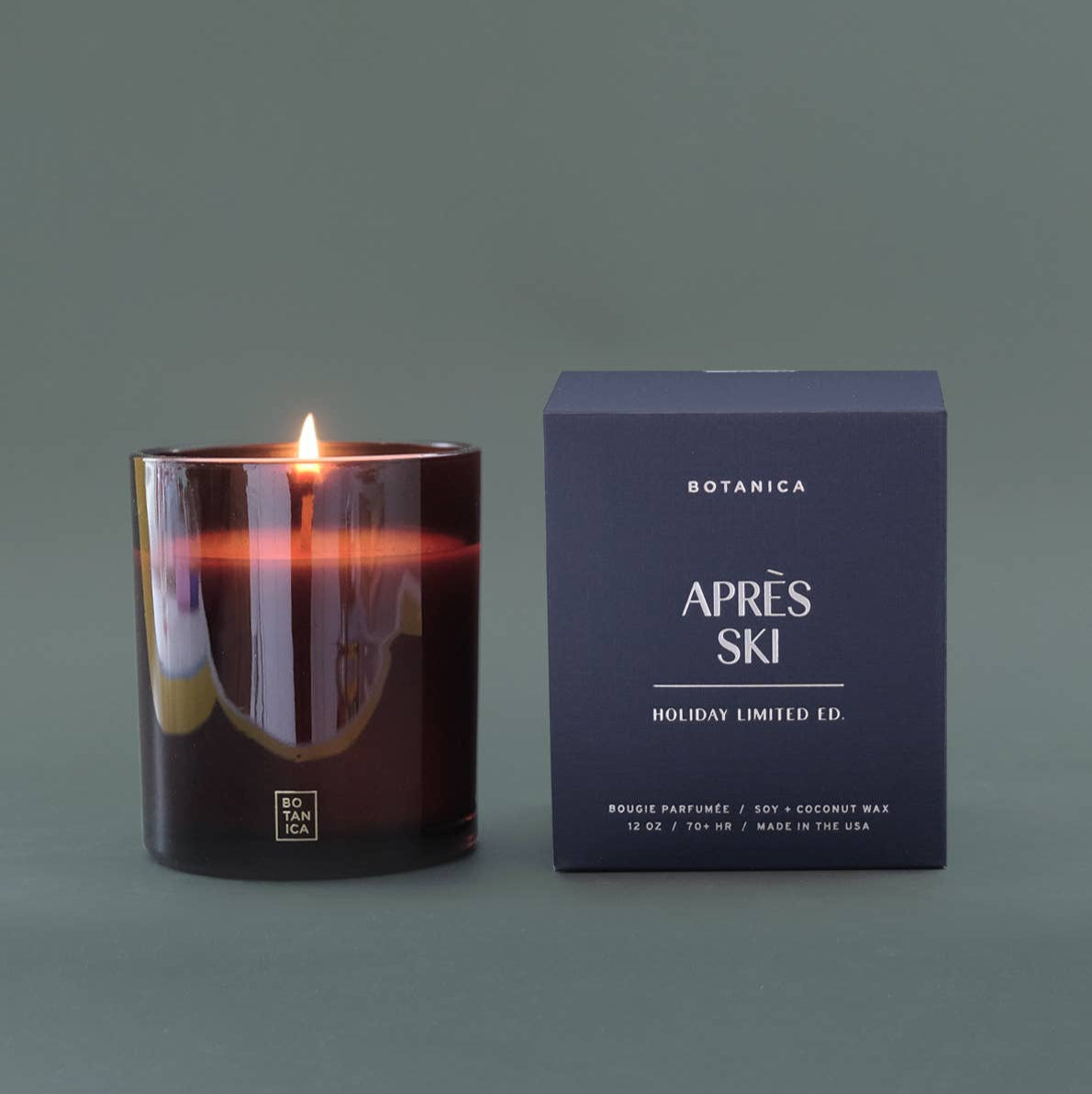 Candle with a lit wick next to its packaging on a dark background