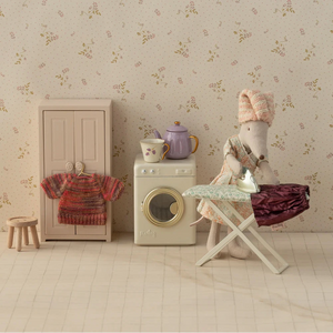 Miniature laundry room setup with a washing machine, dryer, and ironing board against a floral wallpapered wall.
