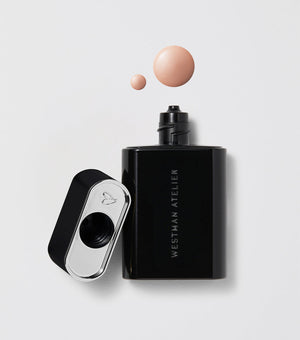 A black bottle of Westman Atelier Liquid Super Loaded Tinted Highlighter with its cap removed, showing a small amount of product headphone on a white background.