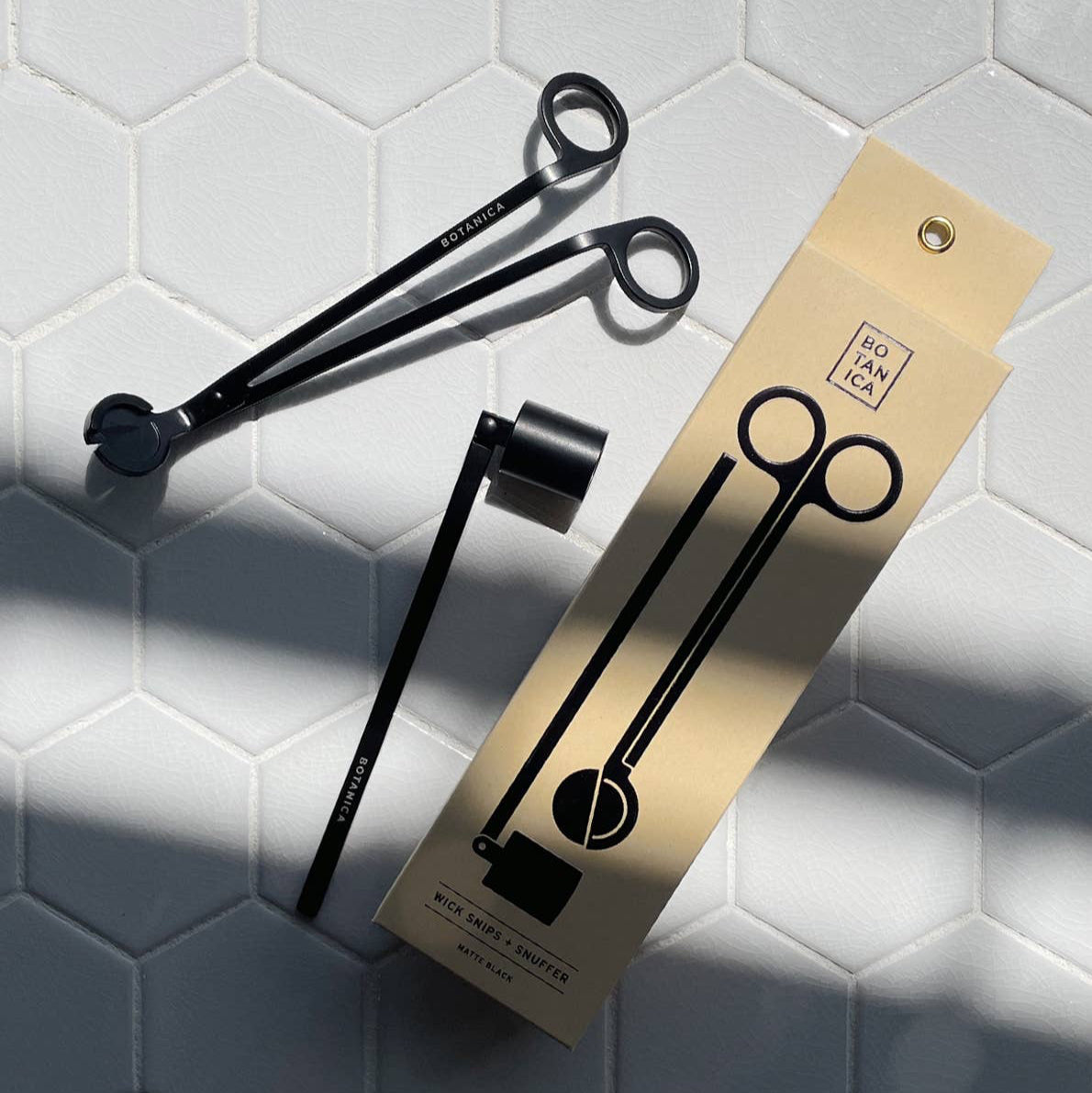 A cream box that says Botanica, next to a candle wick trimmer and snuffer, on a white tile surface. 