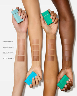 Hands holding green containers with makeup swatches on arms against a white background
