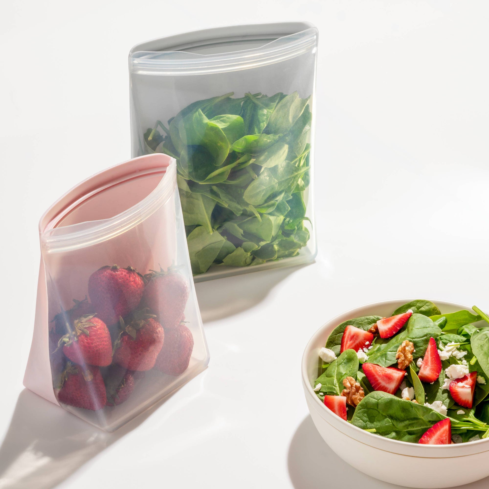 Reusable silicone food storage bags with strawberries and spinach, next to a bowl of salad.