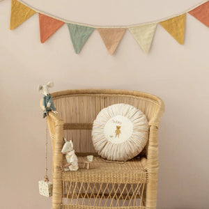 Wicker chair with decorative pillow and plush toys against a neutral background with a colorful triangle banner.