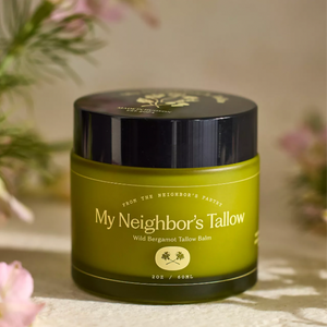 Green jar of 'My Neighbor's Tallow' balm with a black lid on a light surface with flowers in the background.