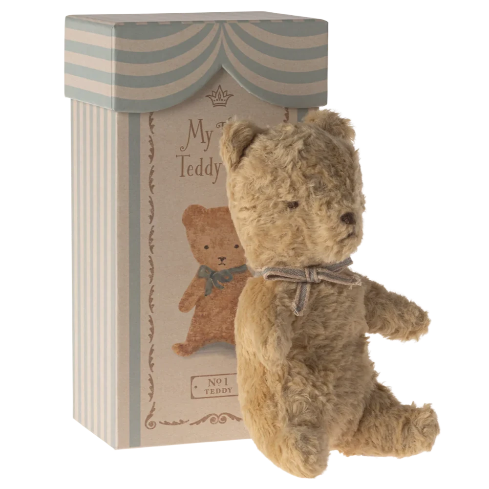 A small, plush teddy bear with a vintage look, sitting next to its packaging box which is also styled with vintage graphics.