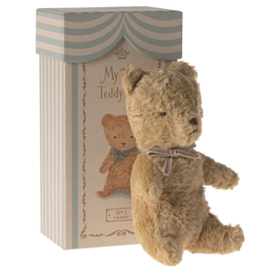 A small, plush teddy bear with a vintage look, sitting next to its packaging box which is also styled with vintage graphics.