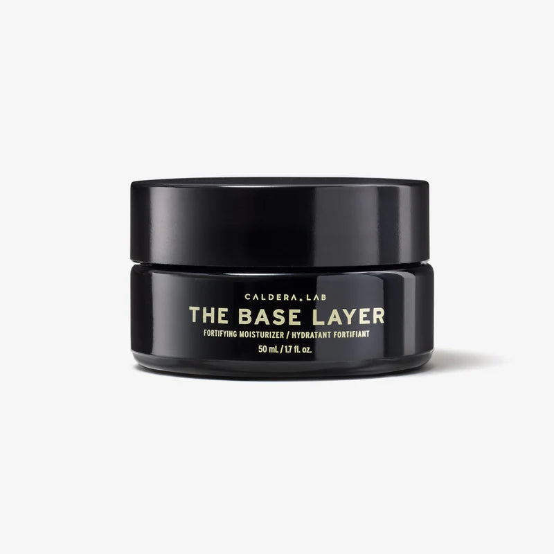 A black jar with a gold and white label reading 'THE BASE LAYER' by Caldera + Lab, indicating a nourishing day moisturizer.