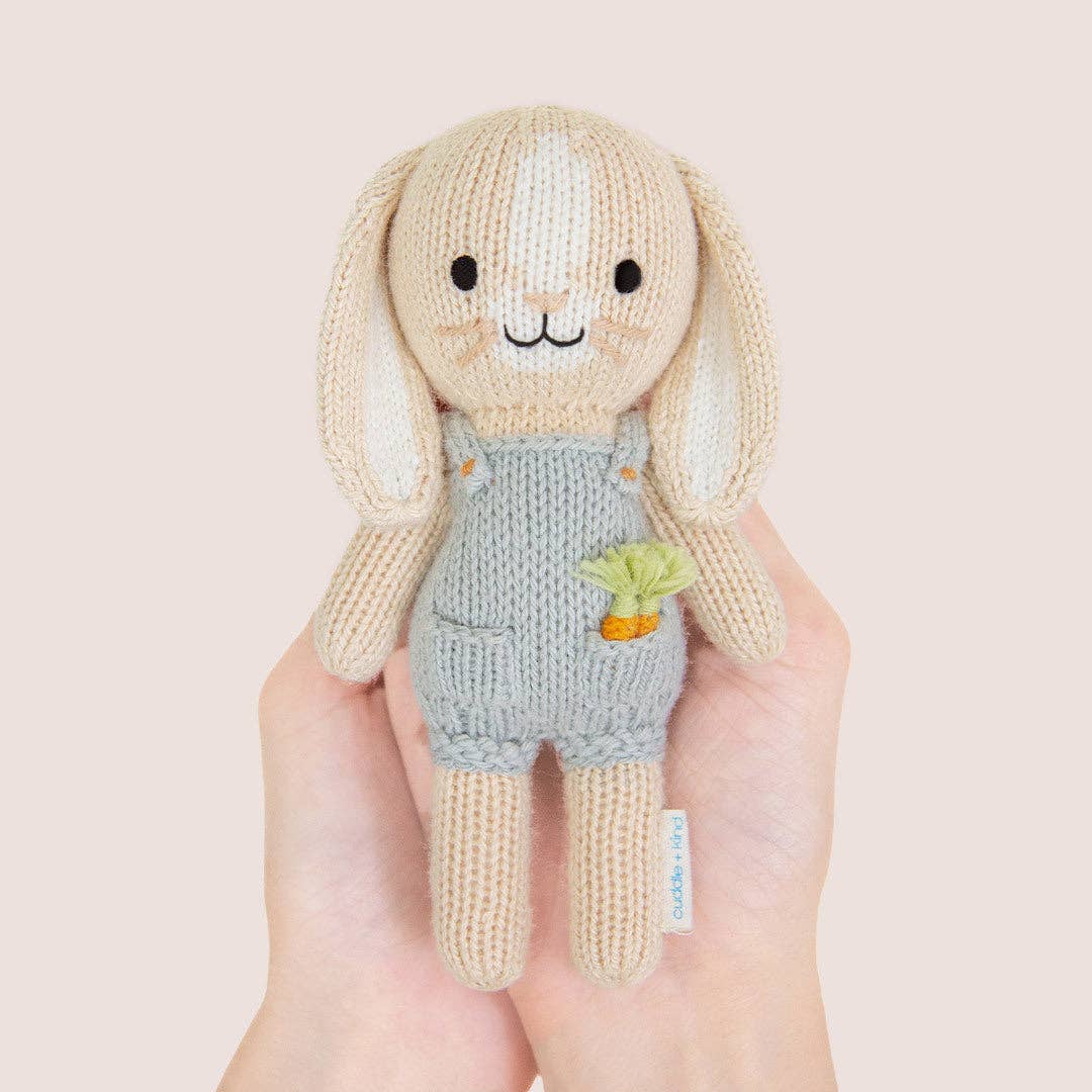 Hands holding a small knit bunny wearing blue overalls with carrots in its pocket. 