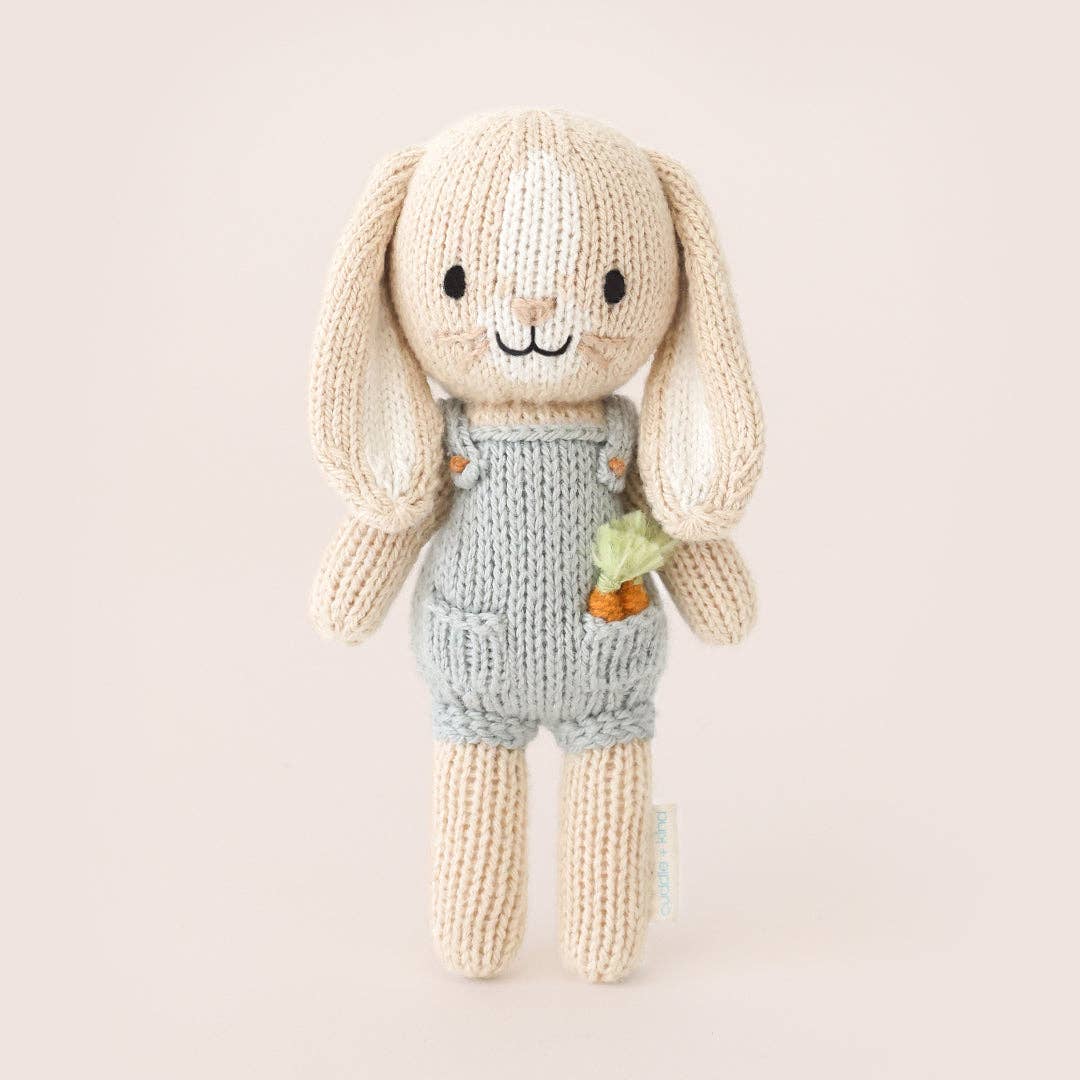 A small knit bunny wearing blue overalls with carrots in its pocket. 