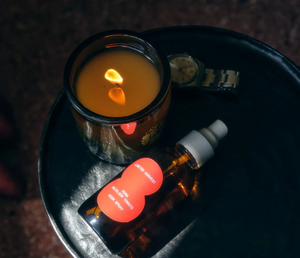 A bottle of tomato room spray, laying next to a lit candle and a watch on a black surface. 