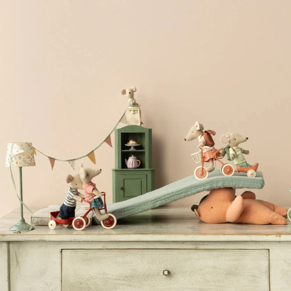 Toy mice on tricycles with a toy bear and furniture in a minimalistic room.