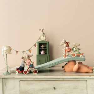 Toy mice on tricycles with a toy bear and furniture in a minimalistic room.