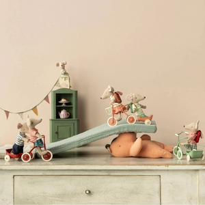 Toy mice on tricycles with a teddy bear and teapot cabinet in a cozy room.
