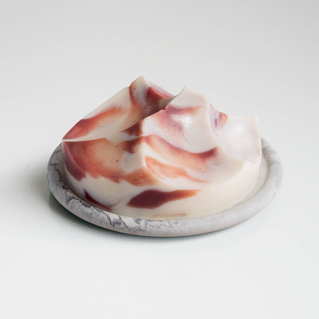 A creamy, swirled bar of soap with a smooth texture, predominantly white with swirls of pink and purple, presented on a grey dish.
