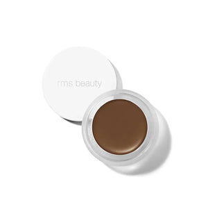 A small glass pan of RMS Beauty UnCoverup Concealer with a white cap that says "RMS Beauty,"