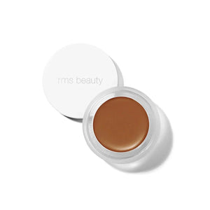 A small glass pan of RMS Beauty UnCoverup Concealer with a white cap that says "RMS Beauty,"