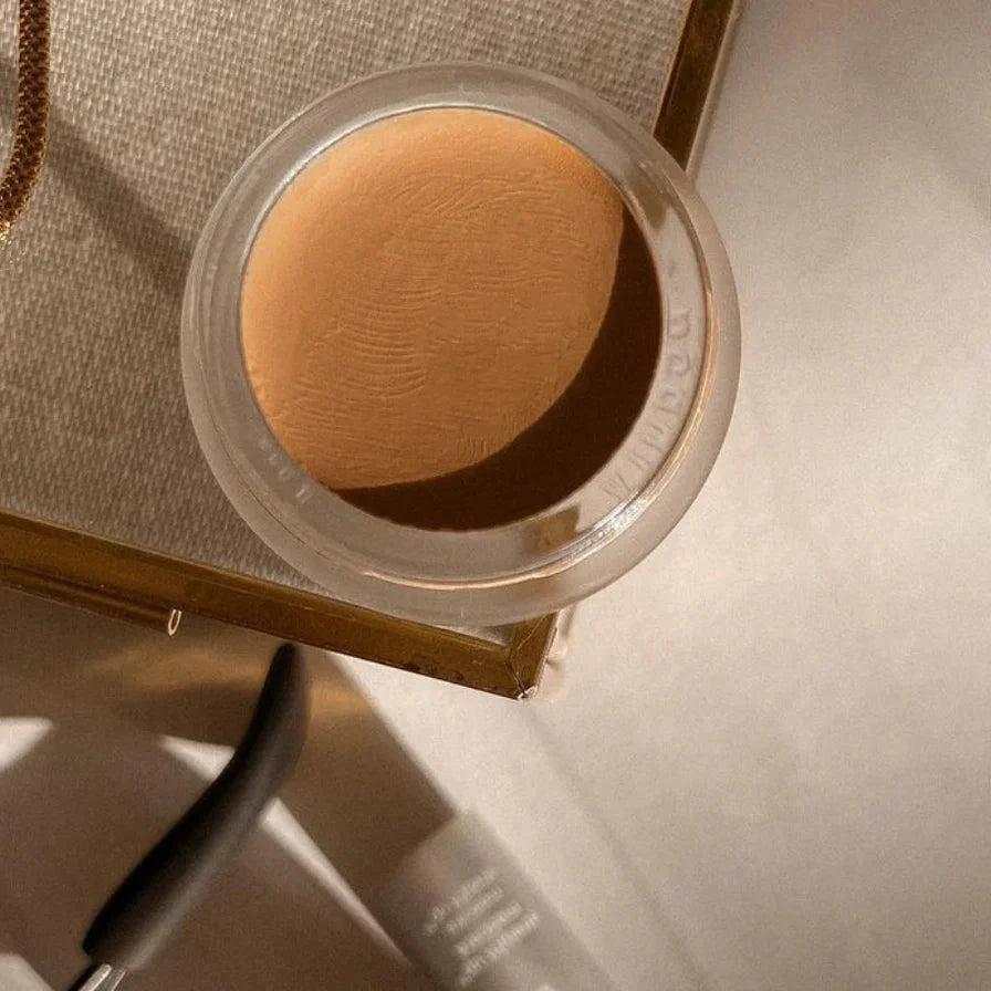 A small glass pan of RMS Beauty UnCoverup Concealer on a beige background. 