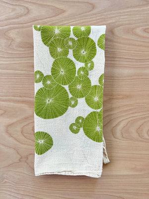 A white cotton tea towel with a green silkscreened urchin pattern, displayed on a wooden surface.